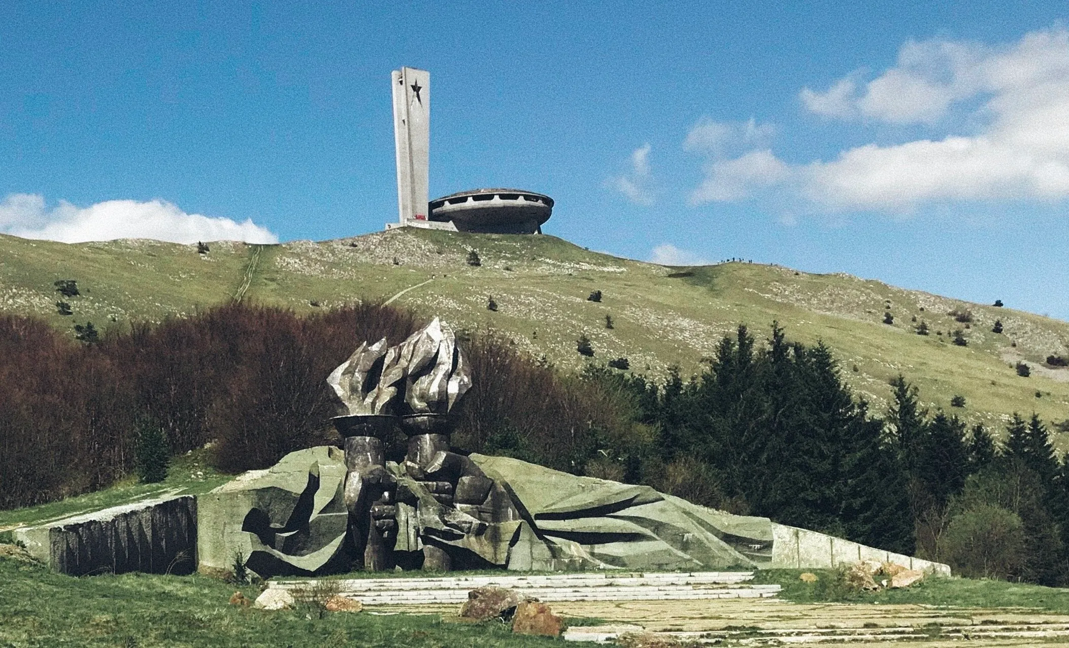 The image depicts the real-life Buzludzha Monument located in Bulgaria. It shows the monument perched atop a grassy hill, with a central tower and a flying saucer-like building with a ring of windows around it. In the foreground, there is a large, abstract metal sculpture featuring dynamic shapes and figures, which seems to be part of a memorial ensemble. The sky is partly cloudy, suggesting a bright yet breezy day. The surrounding landscape is a mix of grassy slopes and forested areas, which adds to the imposing and somewhat isolated setting of the monument. The monument itself has a stark, Brutalist architectural style, standing out against the natural backdrop.